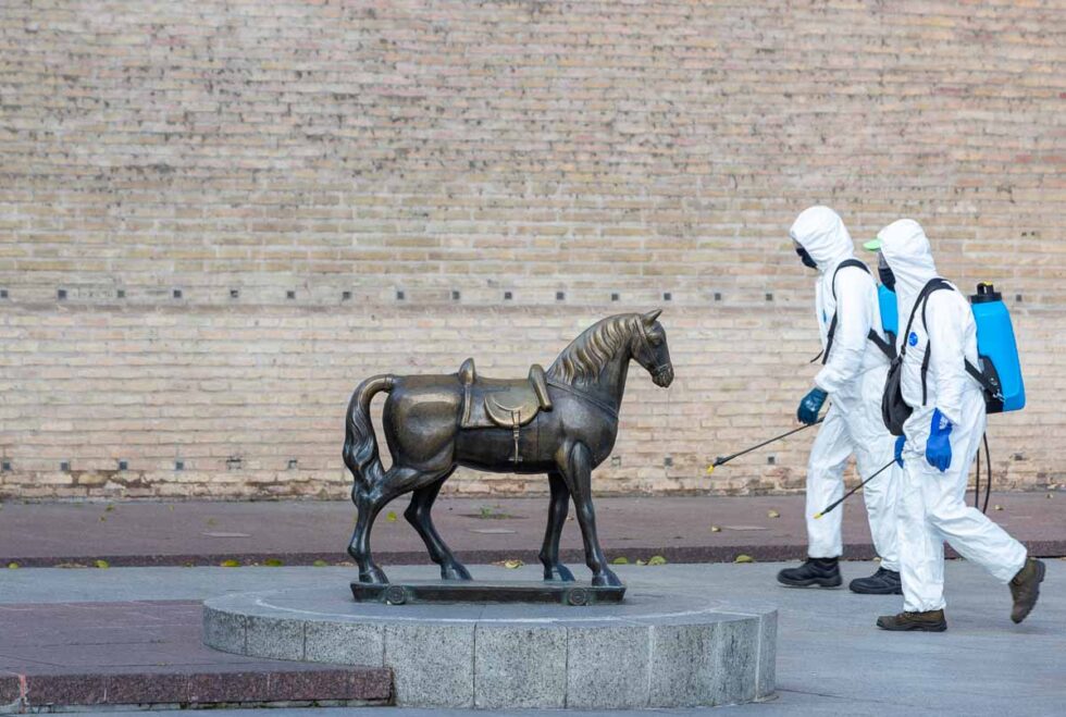 Dos miembros de limpieza equipados con EPIs desinfectan la plaza junto al caballito de La Lonja