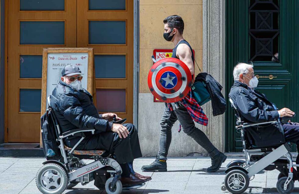 Contraste de la pandemia, un joven con mascarilla pasa con un escudo de Capitan América junto a dos personas en sillas de ruedas