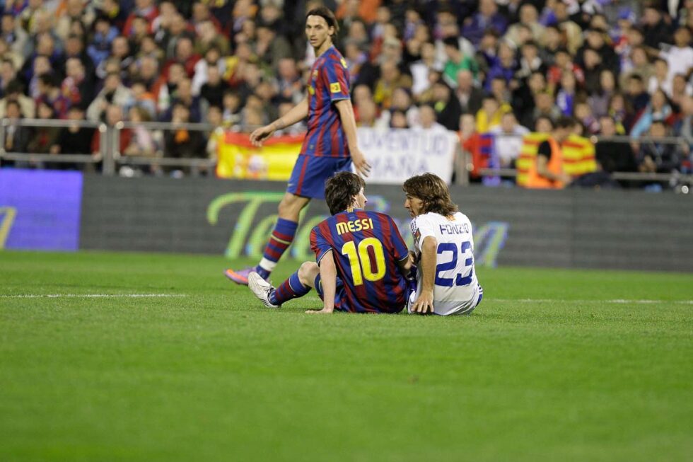 Los jugadores Messi y Leo Ponzio sentados en el campo de fútbol tras una jugada de acción