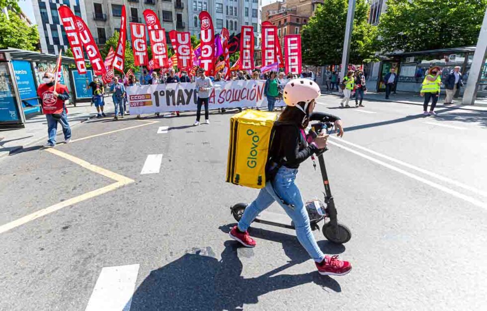 Un repartidor de Glovo pasa delante de la cabeza de la manifestación de UGT y CCOO