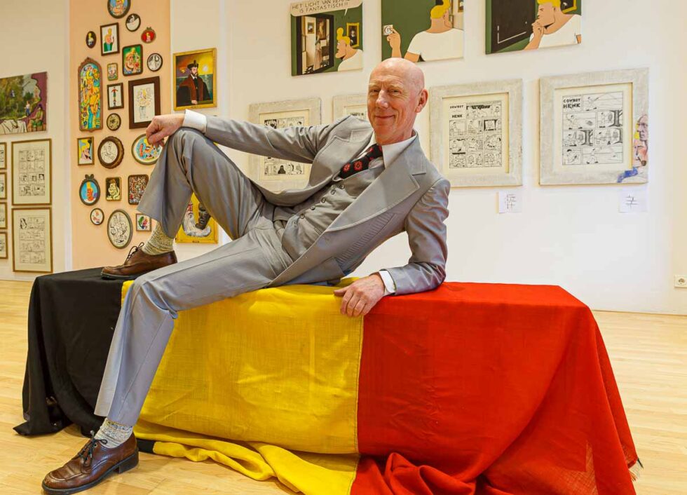 Retrato de Peter Van Heirseele posando sobre una bandera belga en la sala donde se exponen sus obras