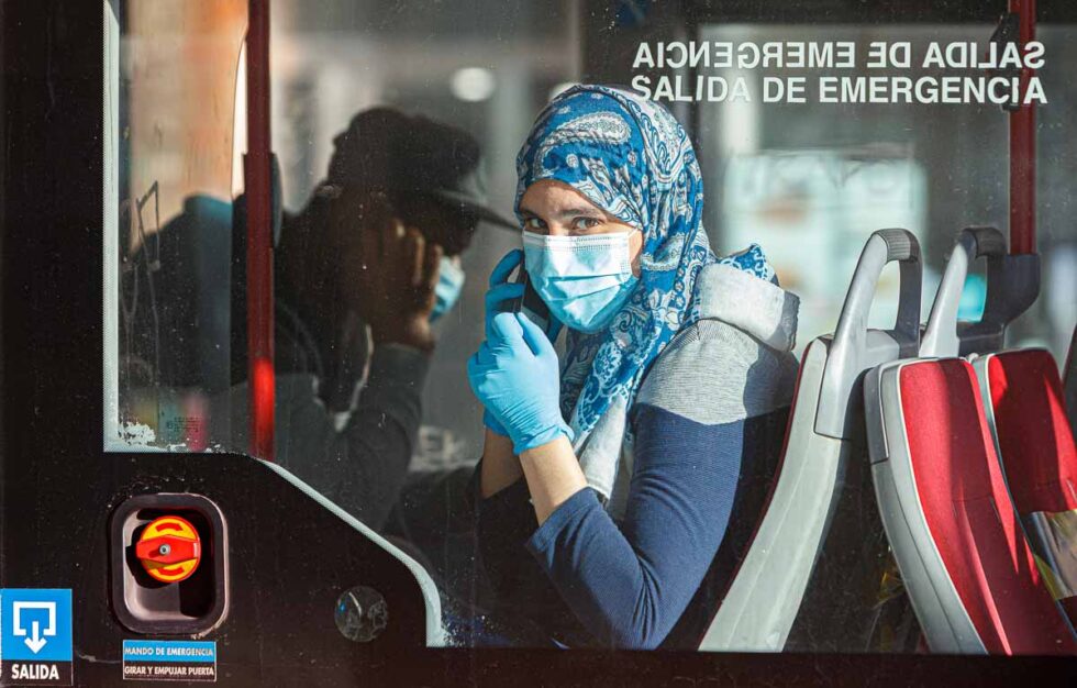 Una mujer con mascarilla y guantes sentada en el interior de un autobús mira a cámara a través del cristal mientras habla por el teléfono móvil